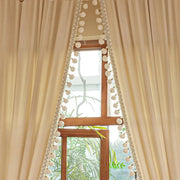 Beige curtains with soft pleats and pom-pom trim framing a tall wooden window