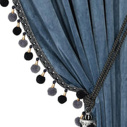 Slate blue velvet curtain with curved black pom-pom trim and decorative tassel tieback