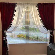 Burgundy velvet curtains with black pom-pom trim layered over white sheer curtains and window blinds