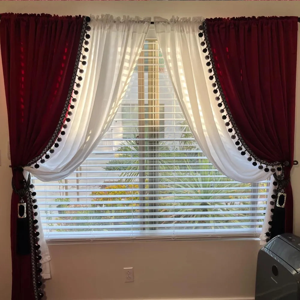 Red velvet curtains with black pom-pom trim and tassel tiebacks layered over white sheer curtains on a bedroom window